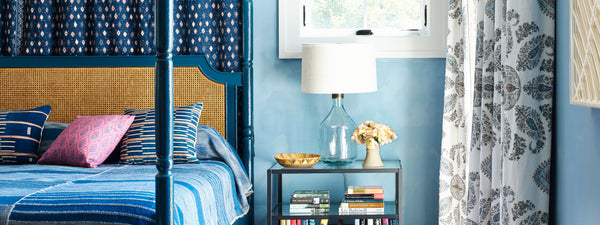 A blue bedroom with a blue painted four poster bed with a cane headboard, a glass table and lamp, and a patterned paisley pair of curtains. the bed is dressed in blue and raspberry colored pillows and a blue alpaca bedcover.