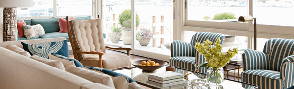 A living room with a sofa, pair of striped chairs, and armchair surrounding a coffee table topped wtih books and yellow flowers. Out the window is the ocean.