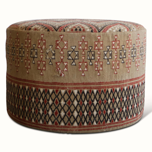 Round upholstered ottoman, beige rug fabric with red and dark blue geometric motifs.