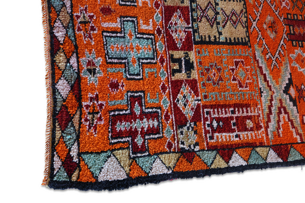 Close-up of orange rug with diamond and cross patterns, dark border, fringed edge